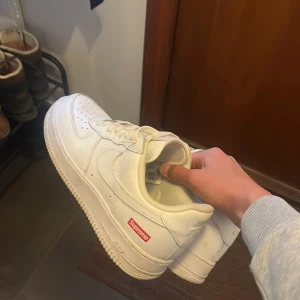 Nike Air Force 1 x Supreme vita sneakers - Nike Air Force 1 x Supreme sneakers i helvitt skinn med klassisk låg siluett. Snygg Supreme-logga i rött på sidan och innersulan. Perforerad tå, snörning och platt sula. Perfekta för dig som gillar clean streetstyle och ikoniska samarbeten.