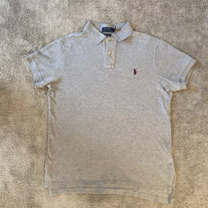 Ralph Lauren piké - Klassisk grå pikétröja från Polo Ralph Lauren med röd broderad logga på bröstet. Tröjan har korta ärmar, krage och två knappar framtill. Tillverkad i mjuk bomull och har en normal passform som sitter snyggt på kroppen. Modellen är 183 cm och nypriset är 1200 kr!