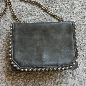 Grå axelväska med kedja Don Donna - Snygg grå axelväska från Don Donna i skinnimitation med coola silverfärgade nitar runt kanterna. Väskan har en kedjerem och flera fack inuti, inklusive ett med dragkedja. Perfekt för att lyfta din outfit med lite edge.