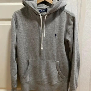 Ralph Lauren hoodie - Säljer en klassisk grå hoodie från Polo Ralph Lauren med broderad logga på bröstet. Tröjan har huva med vita snören och en stor magficka. Perfekt för en avslappnad stil.