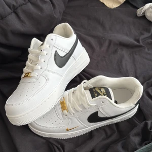 Nike Air Force 1 vit/svart/guld - Snygga Nike Air Force 1 sneakers i vitt skinn med svart swoosh och detaljer i guld. Klassisk siluett med perforerad tå, vit platt sula och snörning. Guldiga detaljer på snörningen och liten guldfärgad swoosh på sidan ger extra lyxig vibe. Säljer för att den var för liten och inte min storlek, byter gärna med en vara med bra skick och i storlek 37,5 eller 38 