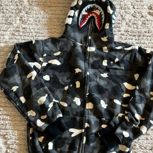 BAPE camo shark hoodie svart/grå - Säljer en ikonisk BAPE shark hoodie med hel dragkedja och camomönster i svart, grått och vitt. Huvan har det klassiska hajansiktet med broderade detaljer och röda accenter. Mjuk bomullskvalitet och ribbade muddar. Perfekt statement-plagg för streetwear. Priset är inte hugget i sten!