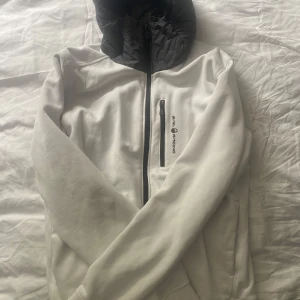 Vit vindjacka från Sail Racing - Snygg vit vindjacka från Sail Racing med svart huva och dragkedja framtill. Jackan har en bröstficka med dragkedja och logga på bröstet. Perfekt för dig som gillar stilren och sportig look. Långärmad och med bekväm passform. 