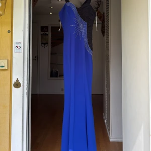 Blå långklänning med strassdetaljer - Snygg blå långklänning med svart överdel och glittrande strassdetaljer i ett stjärnmönster. Klänningen har en axelbandslös design på ena sidan och bred axelrem på andra, vilket ger en cool oneshoulder-look. Perfekt för bal eller festliga tillfällen.