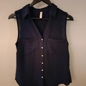 Svart ärmlös blus från Amisu - Svart ärmlös blus från Amisu med klassisk krage och knappar framtill. Toppen har två bröstfickor och är tillverkad i ett lätt och luftigt tyg, perfekt för varmare dagar. Enkel och stilren design som funkar till många olika looks. Aldrig använd.