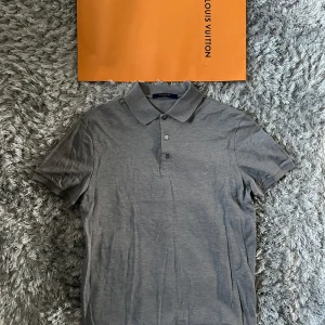 Grå pikétröja från Louis Vuitton - Snygg grå pikétröja från Louis Vuitton med klassisk krage och tre knappar framtill. Diskret LV-logga broderad på bröstet. Tillverkad i bomull och perfekt för en clean och stilren look. Passar dig som gillar exklusiva märken och enkel design.