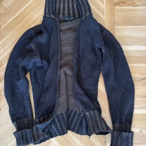 Mörkbrun stickad cardigan från Tiger of Sweden - Mysig mörkbrun stickad cardigan från Tiger of Sweden med breda ribbade muddar. Tröjan har en loose passform och är perfekt för kyliga dagar. Snygga detaljer med ränder på insidan av huvan och muddar.