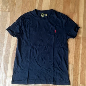 Svart t-shirt från Polo Ralph Lauren - Svart t-shirt från Polo Ralph Lauren med röd broderad logga på bröstet. Modellen är custom slim fit och har rund halsringning och korta ärmar. Tillverkad i mjuk bomull, perfekt för en clean och stilren look.