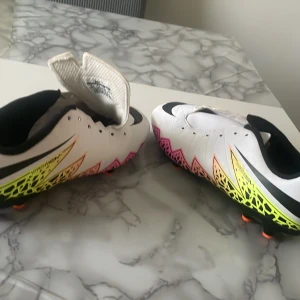 Nike Hypervenom vita fotbollsskor - Nike Hypervenom fotbollsskor i vitt med svarta detaljer och coolt mönster i neonrosa, orange och gult längs sidorna. Skorna har snörning och låg profil, perfekt för dig som vill sticka ut på planen. Snygg design med svart swoosh och mönstrad sula.