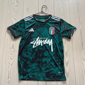 Italien Adidas x Stüssy fotbollströja S - Snygg grön och svart fotbollströja från Adidas i samarbete med Stüssy och Italien. Tröjan har korta ärmar, coolt grafiskt mönster och Italiens klubbmärke på bröstet. Materialet är lätt och andas, perfekt för fotboll eller streetwear.