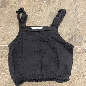 Svart linnetopp från H&M - Svart linnetopp från H&M med breda axelband och coolt strukturerat tyg. Toppen har resår nertill för en avslappnad passform och är perfekt för varma dagar. Enkel och stilren design som funkar till det mesta.