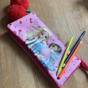 Cherry Bomb rosa plånbok med körsbär - Supergullig rosa plånbok från Cherry Bomb med körsbärsmönster, hjärtan och illustration av två tjejer på framsidan. Tre fack med dragkedja och stora röda fluffiga bollar som dekoration. Perfekt för att hålla koll på småsaker i väskan.