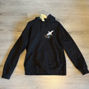 Svart hoodie från Axel Arigato - Svart hoodie från Axel Arigato med broderad vit fågel på bröstet. Klassisk känguruficka framtill, snörning i huvan och ribbade muddar. Perfekt för dig som gillar stilrena streetwear-plagg med en twist. 