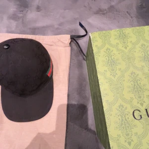 Svart keps från Gucci med GG-mönster - Snygg svart keps från Gucci med klassiskt GG-monogram över hela kepsen och ikoniska gröna och röda ränder på sidan. Justerbar rem baktill och tillverkad i bomull. Perfekt för dig som vill ha en exklusiv och trendig accessoar.