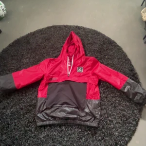 Jordan röd och svart vindjacka med huva - Snygg röd och svart vindjacka från Jordan med huva och vit dragkedja framtill. Jackan har klassisk Jumpman-logga på bröstet och texten 'Air Jordan 23' på magen. Tillverkad i lätt polyester, perfekt för blåsiga dagar. Färgblockad design och elastisk nederkant för skön passform.