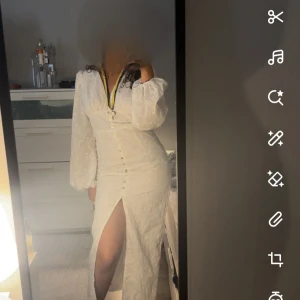 Vit långklänning med slits och puffärm - Supersnygg vit långklänning med v-ringning, puffiga långärmar och markerad midja. Klänningen har knappar framtill och en hög slits som ger en extra touch. Broderade blommor i tyget och romantisk känsla. Perfekt för dig som vill sticka ut!