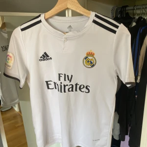 Real Madrid Sergio Ramos matchtröja - Vit Real Madrid fotbollströja från Adidas med svarta detaljer, klubbmärke och Fly Emirates-tryck på bröstet. La Liga-märke på ärmen och Sergio Ramos #4 på ryggen. Tillverkad i lätt och ventilerande material (Climacool). Perfekt för dig som älskar fotboll och Real. (Barntröja)