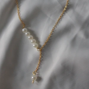 Guldhalsband med pärlor - Ett elegant halsband med tunn guldfärgad kedja och vita pärlor i rad. Kedjan har en asymmetrisk design där en ensam pärla hänger ned från ena sidan. Perfekt för dig som gillar stilrena och trendiga smycken med en touch av lyx.