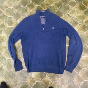 Blå tröja med dragkedja från Gant - Säljer en snygg blå tröja från Gant med halv dragkedja och broderad logga på bröstet. Perfekt för kyligare dagar med sin stilrena design och bekväma passform.