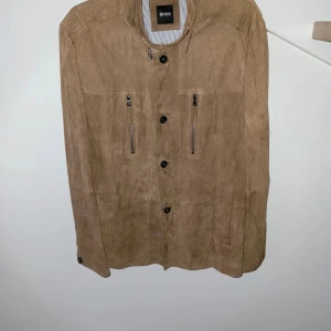Beige mockajacka från Hugo Boss - Säljer en snygg beige mockajacka från Hugo Boss. Jackan har långa ärmar och stängs med knappar. Den har två dragkedjefickor på bröstet och en stilren design. Perfekt för en stilmedveten look. En knapp hänger lite löst men det är enkelt att åtgärda. 