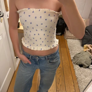 Blommig axelbandslös topp - Säljer en söt axelbandslös topp med små blå blommor på en vit bakgrund. Toppen är croppad och har en volangdetalj vid nederkanten. Perfekt för sommarens varma dagar!