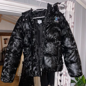 Svart camo pufferjacka från Juicy Couture - Säljer en svart pufferjacka från Juicy Couture med ett coolt camouflagemönster. Jackan har en dragkedja framtill och en mysig huva. Perfekt för kyliga dagar när du vill hålla stilen. 🖤