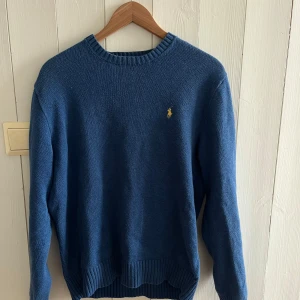 Blå stickad tröja från Ralph Lauren - Säljer en snygg blå stickad tröja från Ralph Lauren med det klassiska logotypbroderiet på bröstet. Tröjan har långa ärmar och ribbade kanter vid hals, ärmslut och nederkant. Perfekt för en stilren look.