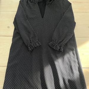 Svart prickig klänning - Snygg svart klänning med vita prickar och v-ringad hals. Den har långa ärmar och en elegant look. Perfekt för att sticka ut med stil.