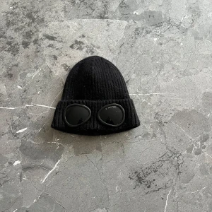 Svart mössa från C.P. Company - Snygg svart mössa från C.P. Company med ribbad design och inbyggda glasögon. Perfekt för att ge din outfit en unik touch. Tillverkad i ett mjukt och bekvämt material.