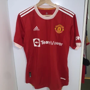 Röd Manchester United fotbollströja från Adidas Rashford  - Säljer en röd Manchester United fotbollströja från Adidas med korta ärmar. Tröjan har vita detaljer och klubbens emblem på bröstet. Perfekt för fans som vill visa sitt stöd. Tillverkad med Heat.Rdy-teknologi för optimal komfort. Rashford på ryggen. Tröjan är äkta