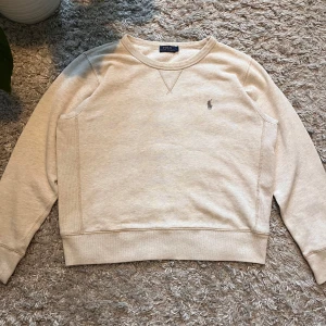 Beige tröja från Ralph Lauren - Säljer en stilren beige tröja från Ralph Lauren med deras ikoniska logga på bröstet. Tröjan har långa ärmar och en klassisk rund halsringning. Perfekt för en avslappnad och snygg look. En fläck på vardera arm, men de är väldigt små och diskreta.