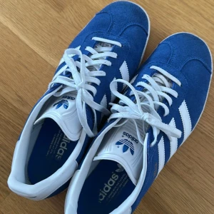 Blå Adidas Gazelle sneakers - Snygga blå Adidas Gazelle sneakers med vita detaljer och snörning. Skorna har en klassisk design med tre vita ränder på sidan och en vit sula. Perfekta för en sportig och stilren look.