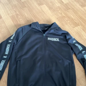 Svart hoodie från Hoodrich - Säljer en svart jacka från Hoodrich med dragkedja framtill. Jackan har långa ärmar med Hoodrich-logga längs ärmarna och på bröstet. Perfekt för en stilren look.