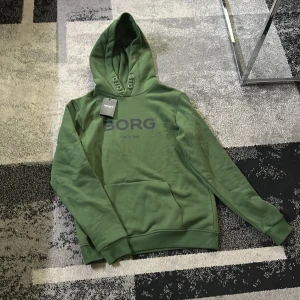 Grön hoodie från Björn Borg - Säljer en snygg grön hoodie från Björn Borg med tryck framtill. Den har en klassisk design med huva och långa ärmar. Perfekt för en avslappnad stil.