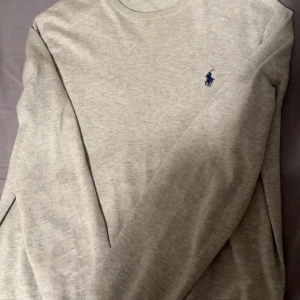 Grå tröja från Polo Ralph Lauren - Säljer en stilren grå tröja från Polo Ralph Lauren med det klassiska logotypbroderiet i blått på bröstet. Tröjan har långa ärmar och en rund halsringning, Bra skick inte alls mycket använd