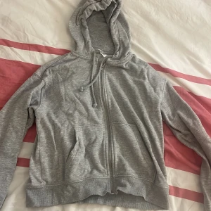 Grå hoodie  - Säljer en klassisk grå hoodie med dragkedja och justerbar huva. Perfekt för en avslappnad stil. Den har långa ärmar och praktiska fickor framtill. Passar perfekt för kyliga dagar eller mysiga hemmakvällar.
