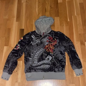 Ed Hardy Hoodie cool - Säljer en cool svart och grå hoodie från Ed Hardy med drak- och rosdesign. Tröjan har en dragkedja framtill och en stor huva. Perfekt för dig som gillar unika och iögonfallande mönster. 🐉🌹