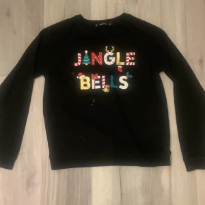Svart jultröja med Jingle Bells-tryck - Säljer en svart tröja med färgglatt 'Jingle Bells'-tryck. Perfekt för julfirandet med roliga detaljer som julgran och renhorn. Tröjan har långa ärmar och är gjord i ett mjukt material. *har fläckar se bild. Kanske går bort