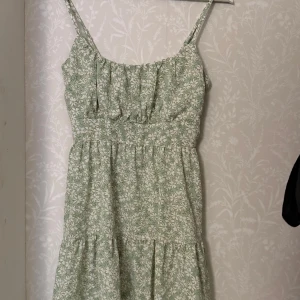 Grön blommig klänning - Söt grön klänning med blommigt mönster och smala axelband. Perfekt för varma dagar med sin lätta och luftiga design. Klänningen har en smickrande passform och är idealisk för sommarens alla tillfällen.