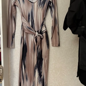 Mönstrad långklänning med knyt - Snygg långklänning med ett unikt mönster i beige och mörkblå toner. Klänningen har långa ärmar och en v-ringad hals. Den har en knytning i midjan som ger en fin siluett. Perfekt för en stilfull look.