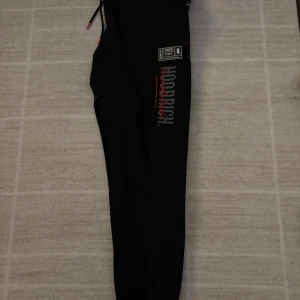 Svarta mjukisbyxor från Hoodrich - Snygga svarta mjukisbyxor från Hoodrich med tryck i vitt och rött. Byxorna har en elastisk midja med snörning och muddar vid bensluten. Perfekta för en avslappnad stil. Riktigt bekväma och snygga.