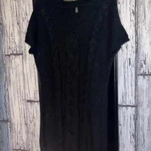 Svart stickad klänning från b.young - Snygg svart stickad klänning från b.young med korta ärmar och flätstickat mönster framtill. Perfekt för kyligare dagar med en mysig och stilren look. Passar bra med leggings eller jeans.