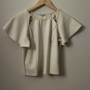 Beige blus med volangärmar - Säljer en stilren beige blus med volangärmar. Perfekt för en elegant look. Blusen har en lös passform och är tillverkad i ett mjukt material.