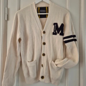 Vit kofta från Morris - Säljer en stilren vit cardigan från Morris i 100% bomull. Cardiganen har knappar framtill, två fickor och ett stort 'M' broderat på bröstet. Den har också svarta ränder på ena ärmen och ribbade muddar. Tröjan är använd 5 gånger och är i mycket bra skick. Nypris ligger på 1699:-