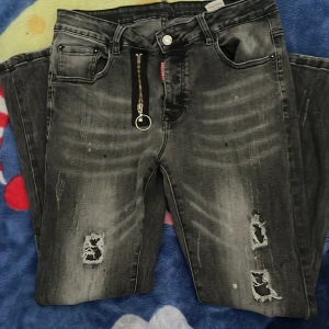 Svarta jeans med slitningar och dragkedja - Coola svarta jeans med slitningar och en unik dragkedja framtill. De har en modern tvättad look med ljusare partier och är perfekta för en avslappnad stil. Passar bra till sneakers eller kängor.