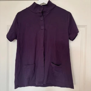 Lila kortärmad tröja med knappar - Säljer en lila kortärmad tröja med hög krage och tre stora knappar framtill. Tröjan har två praktiska fickor på framsidan. Perfekt för en avslappnad stil.
