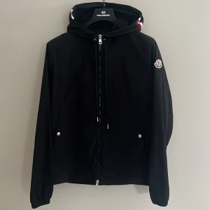 Svart jacka från Moncler - Snygg svart jacka från Moncler med dragkedja och huva. Jackan har långa ärmar och ett Moncler-märke på ärmen. Perfekt för en stilren look nfc funkar 