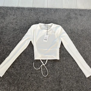 Vit ribbad topp från NA-KD - Säljer en stilren vit ribbad topp från NA-KD. Toppen har långa ärmar och en knytning i midjan som ger en snygg detalj. Perfekt för att skapa en trendig look. Liten i storlek.