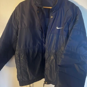 Blå pufferjacka från Nike - Säljer en blå pufferjacka från Nike med dragkedja och tryckknappar. Jackan har långa ärmar och en stilren design med Nike-logga på bröstet och ryggen. Perfekt för kyligare dagar. Strl M