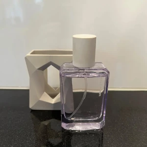 Parfym från Ariana Grande - Snygg parfymflaska med en modern design från Ariana Grande. Flaskan är genomskinlig med en ljuslila ton och har en vit kork. Den kommer med en unik geometrisk hållare i ljusgrått som ger en stilren look.